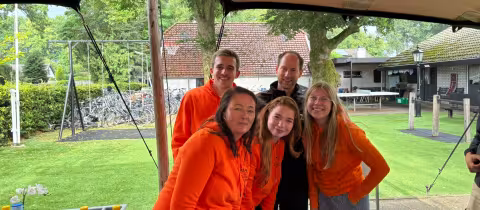 Vijf kampbegeleiders poseren lachend in oranje truien onder de tent.
