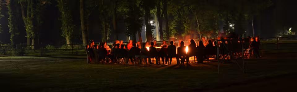 Kinderen zitten samen rond het kampvuur onder hoge bomen.