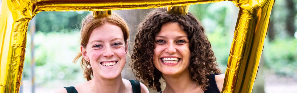 Twee lachende kampleidsters poseren achter een gouden ballonlijst
