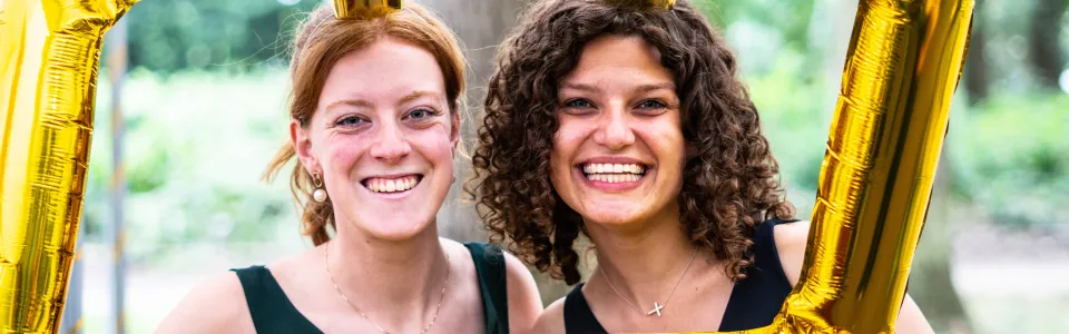 Twee lachende kampleidsters poseren achter een gouden ballonlijst