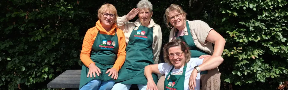 Vier vrijwilligsters poseren vrolijk in groene kampschorten bij een picknickbank.