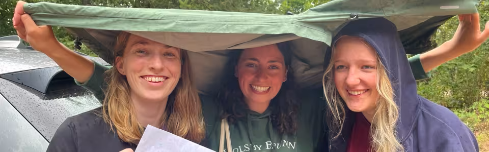 Drie lachende kampleidsters schuilen samen onder een tentzeil in de regen