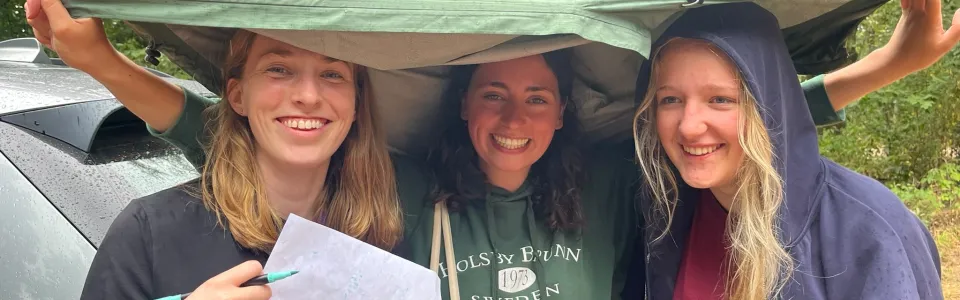 Drie lachende kampleidsters schuilen samen onder een tentzeil in de regen