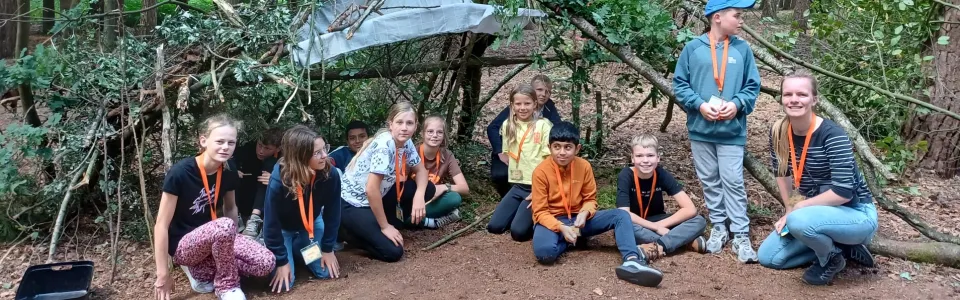Kinderen en leiding poseren trots bij hun zelfgebouwde boshut
