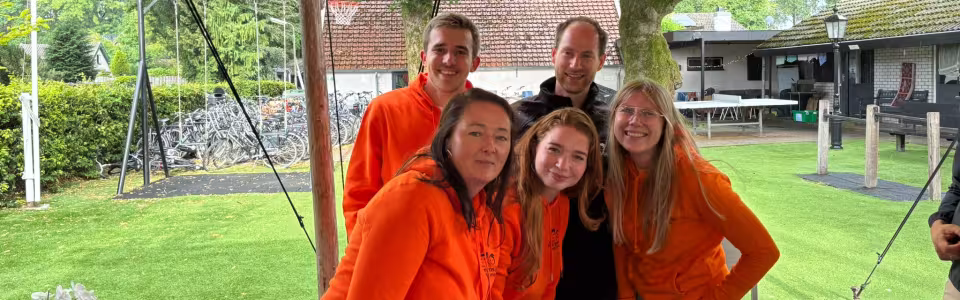 Vijf kampbegeleiders poseren lachend in oranje truien onder de tent.